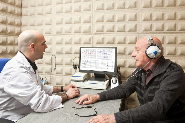 Anziano con cuffie audiometriche bianche e uomo in camice davanti a un monitor con audiogrammi, in cabina fonoassorbente